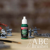 Bottle of the Army Painter Fanatics in the colour Guardian Green with various wargaming models and an ABC paintbrush in the background, on a wooden surface.