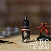 Bottle of the Army Painter Fanatics Metallic paint in the colour Gun Metal with various wargaming models and an ABC paintbrush in the background, on a wooden surface.