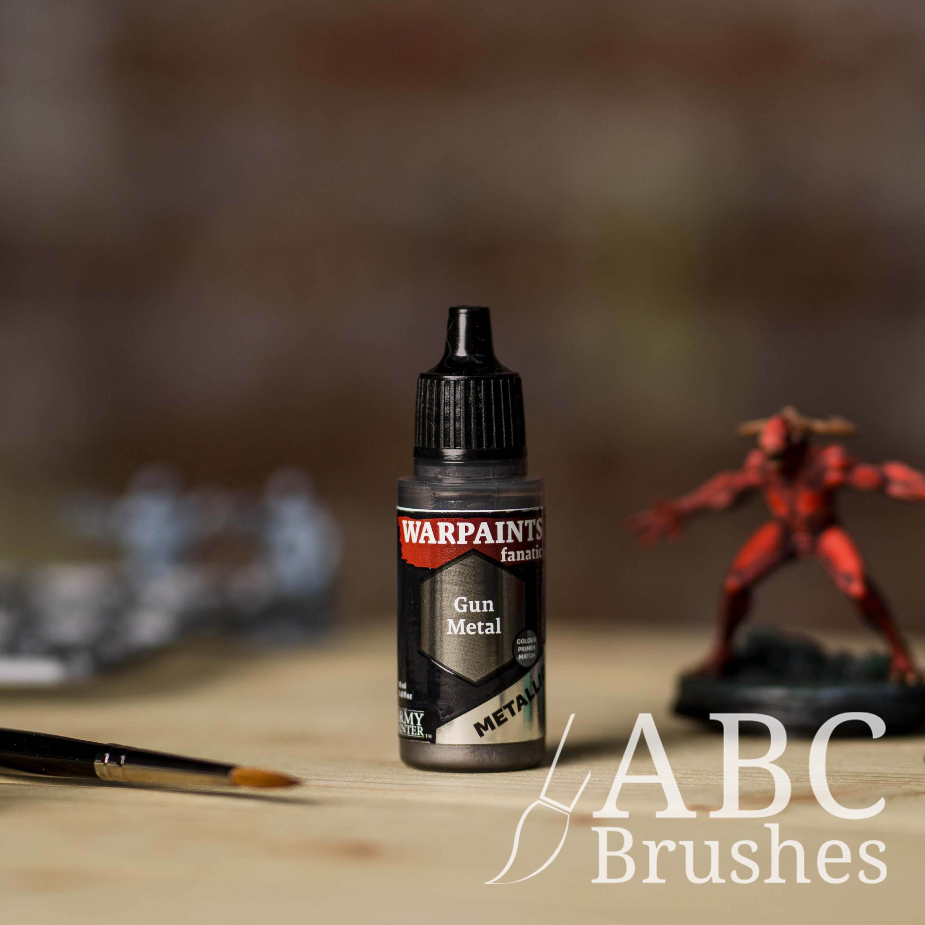 Bottle of the Army Painter Fanatics Metallic paint in the colour Gun Metal with various wargaming models and an ABC paintbrush in the background, on a wooden surface.