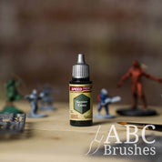 Bottle of the Army Painters Speedpaint in the colour Gunner Camo, with wargaming models and an ABC paintbrush in the background, on a wooden surface.