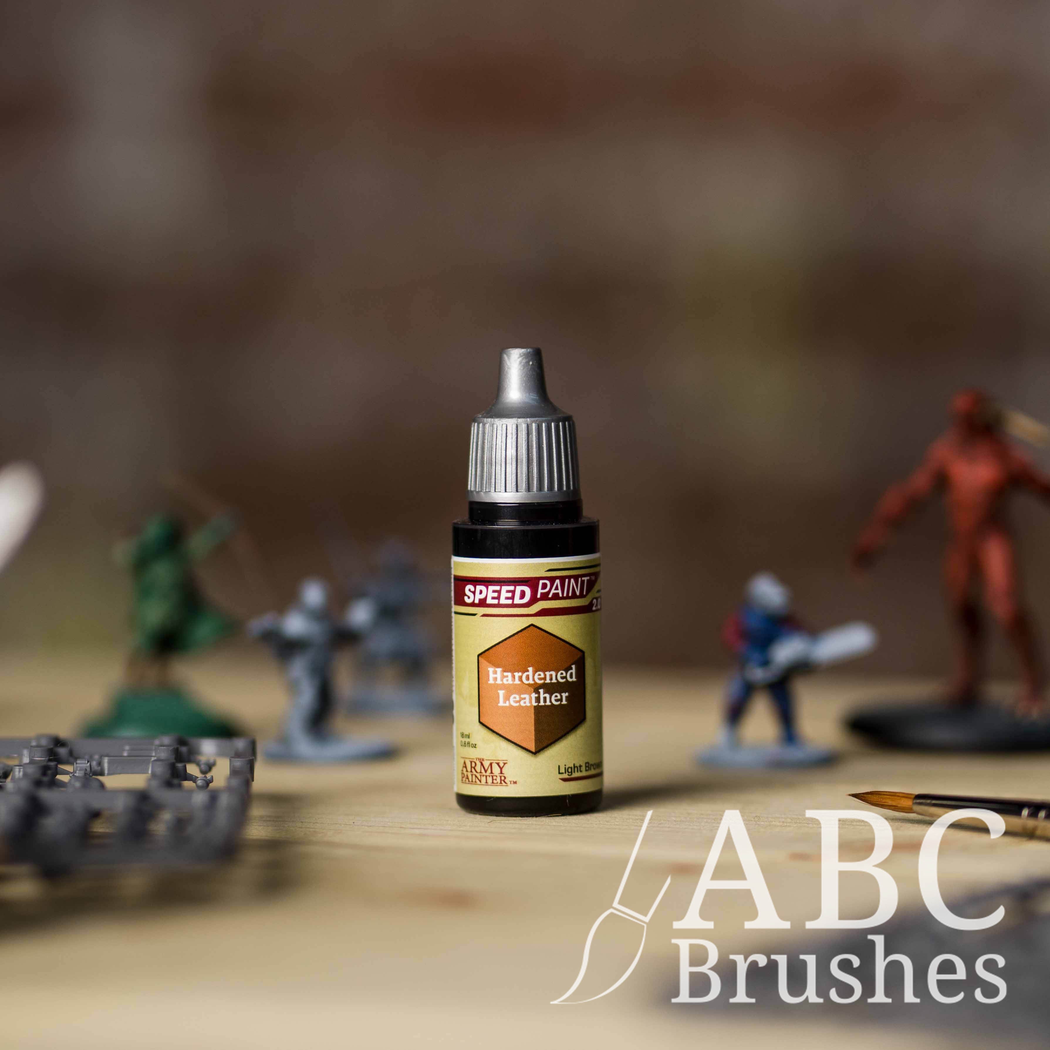 Bottle of the Army Painters Speedpaint in the colour Hardened Leather, with wargaming models and an ABC paintbrush in the background, on a wooden surface.