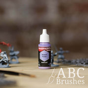 Bottle of the Army Painter Fanatics in the colour Hexed Violet with various wargaming models and an ABC paintbrush in the background, on a wooden surface.