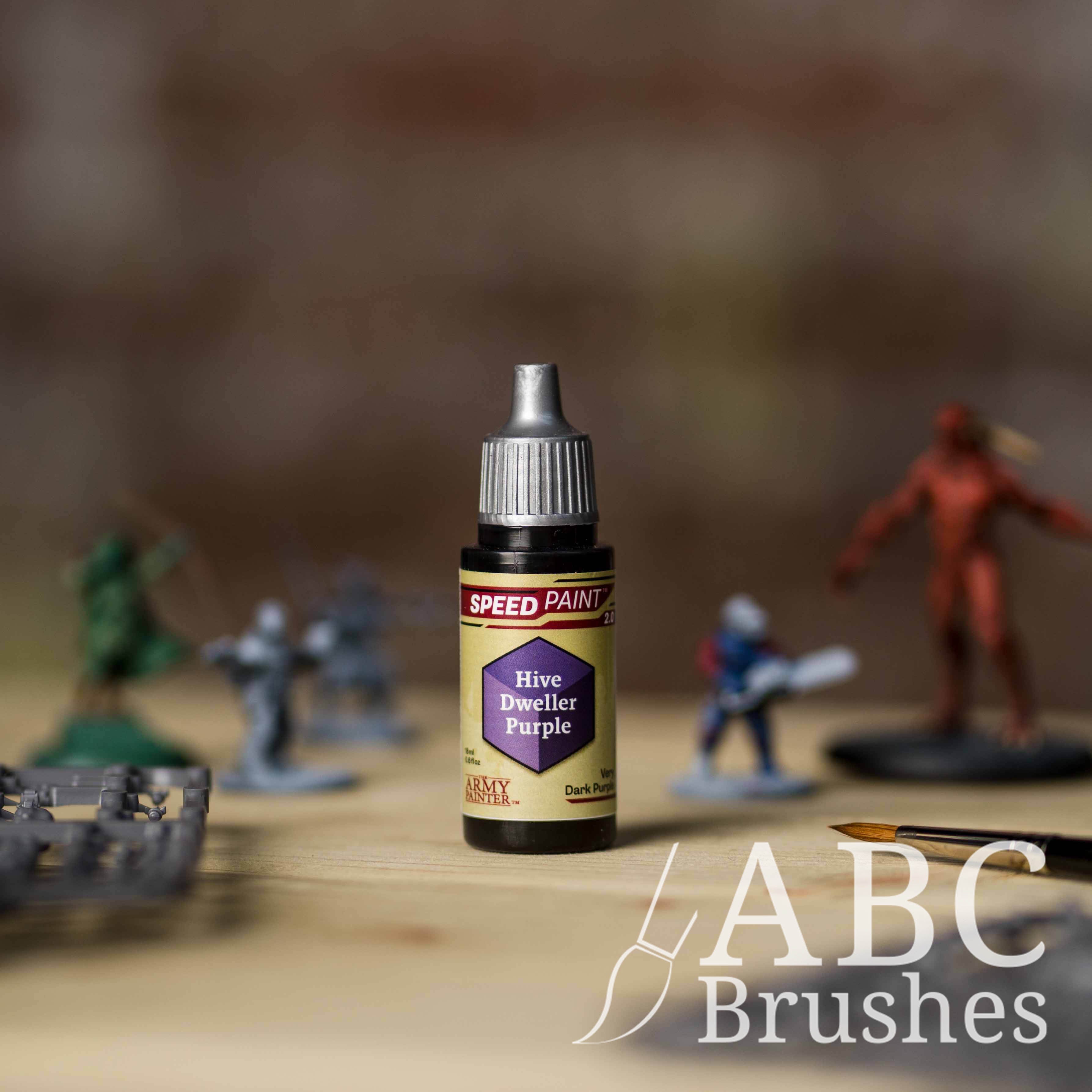 Bottle of the Army Painters Speedpaint in the colour Hive Dweller Purple, with wargaming models and an ABC paintbrush in the background, on a wooden surface.