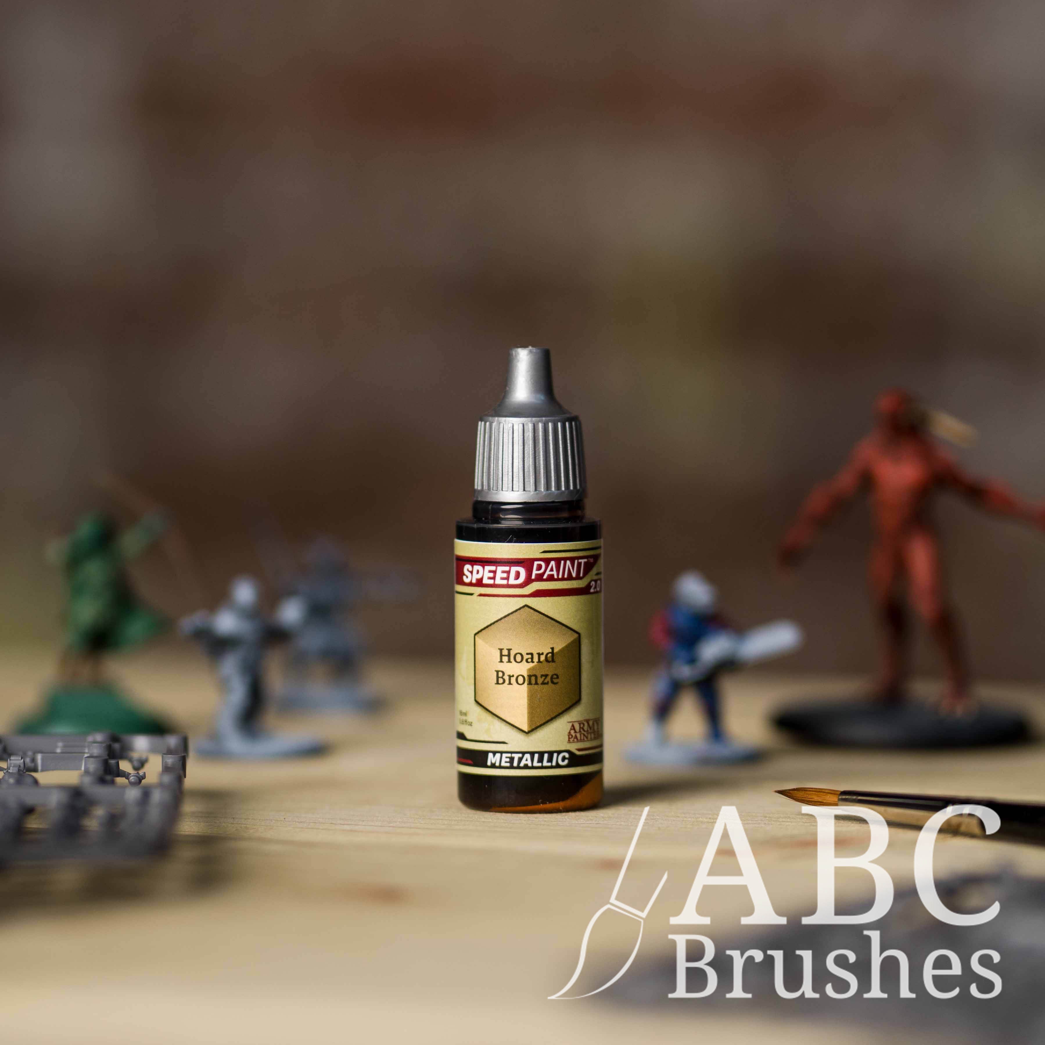 Bottle of the Army Painters Metallic Speedpaint in the colour Hoard Bronze, with wargaming models and an ABC paintbrush in the background, on a wooden surface.