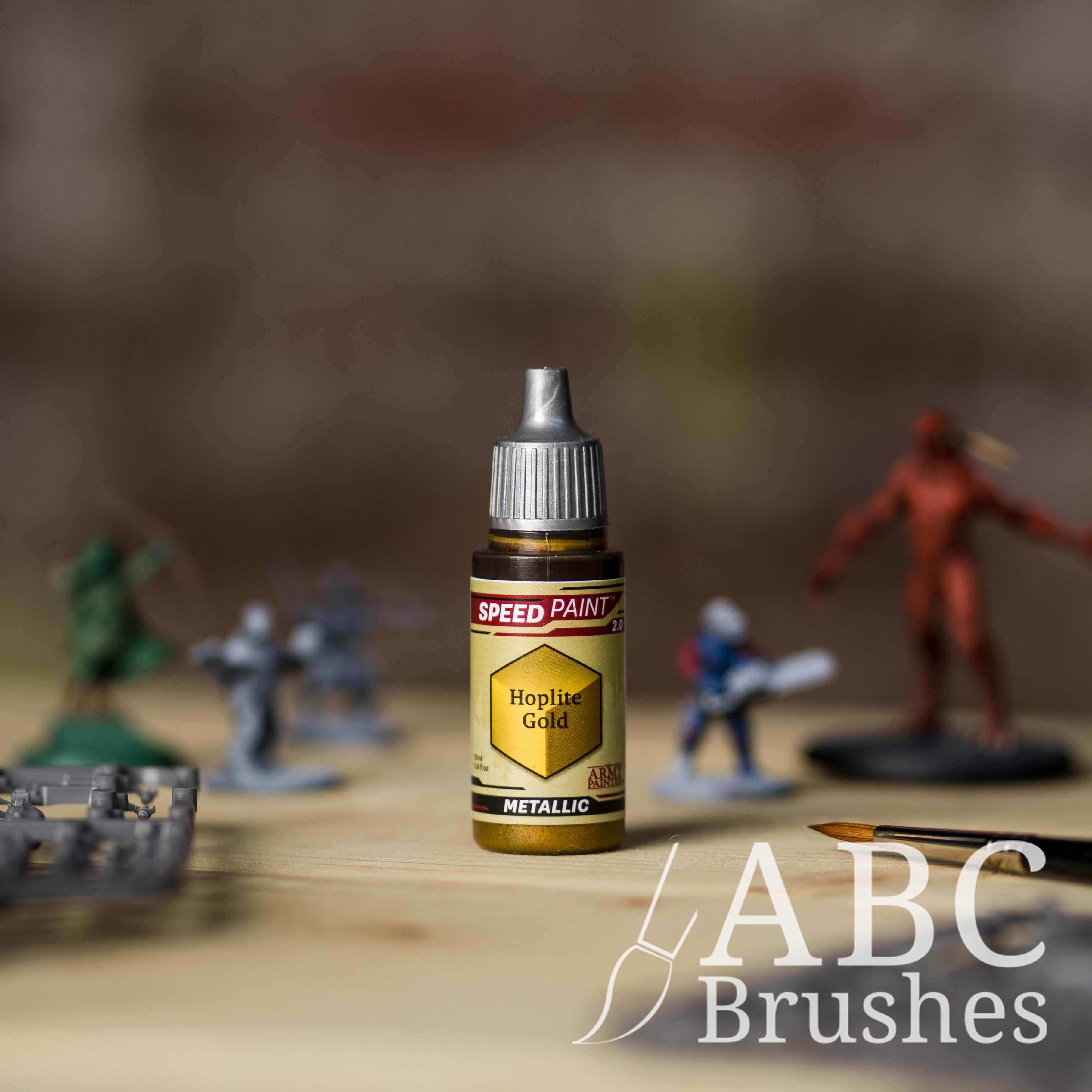 Bottle of the Army Painters Metallic Speedpaint in the colour Hoplite Gold, with wargaming models and an ABC paintbrush in the background, on a wooden surface.