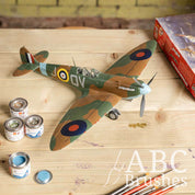 Scale model plane painted in green and brown camouflage, next to tins of Humbrol Enamel paint, brushes and a model box on a wooden table. 