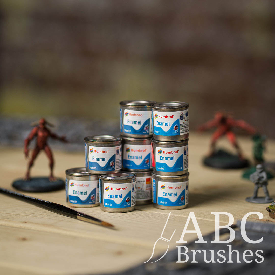 Tins of Humbrol Enamel paint stacked on top of each other on a wooden table, next to miniatures and sprues and a paint brush. 