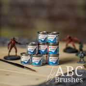Tins of Humbrol Enamel paint stacked on top of each other on a wooden table, next to miniatures and sprues and a paint brush. 