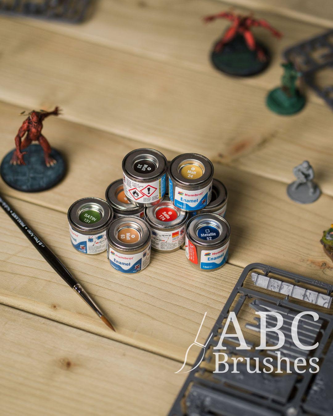 Collection of enamel paint cans on a wooden surface with minis and various miniature painting tools.