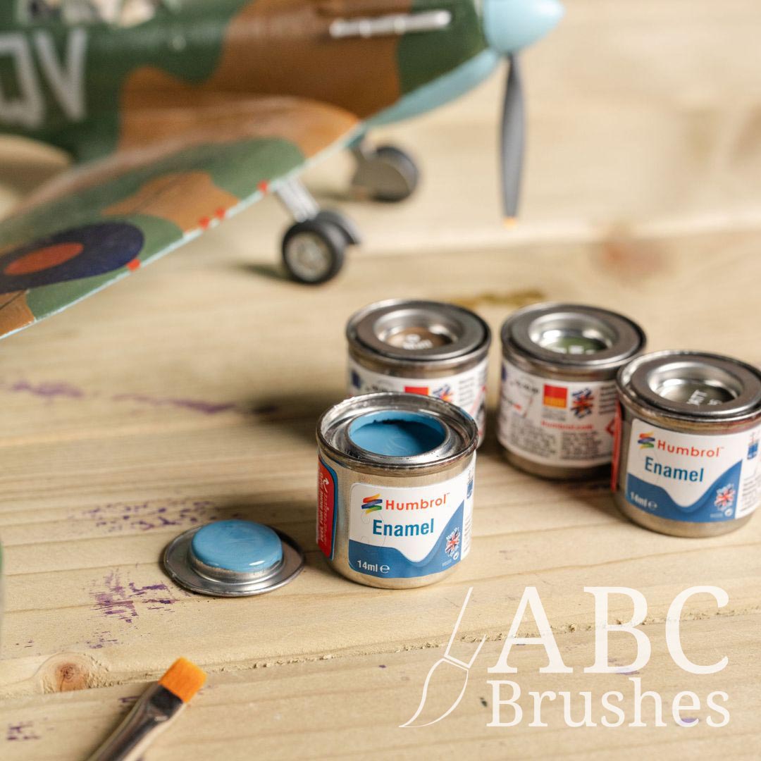 Four cans of humbrol enamel paints, next to a scale model plane and flat paint brush, on a wooden surface.  