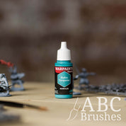 Bottle of the Army Painter Fanatics in the colour Hydra Turquoise with various wargaming models and an ABC paintbrush in the background, on a wooden surface.
