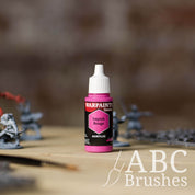 Bottle of the Army Painter Fanatics in the colour Impish Rouge with various wargaming models and an ABC paintbrush in the background, on a wooden surface.