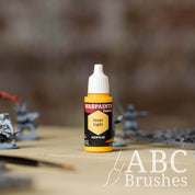 Bottle of the Army Painter Fanatics in the colour Inner Light with various wargaming models and an ABC paintbrush in the background, on a wooden surface.