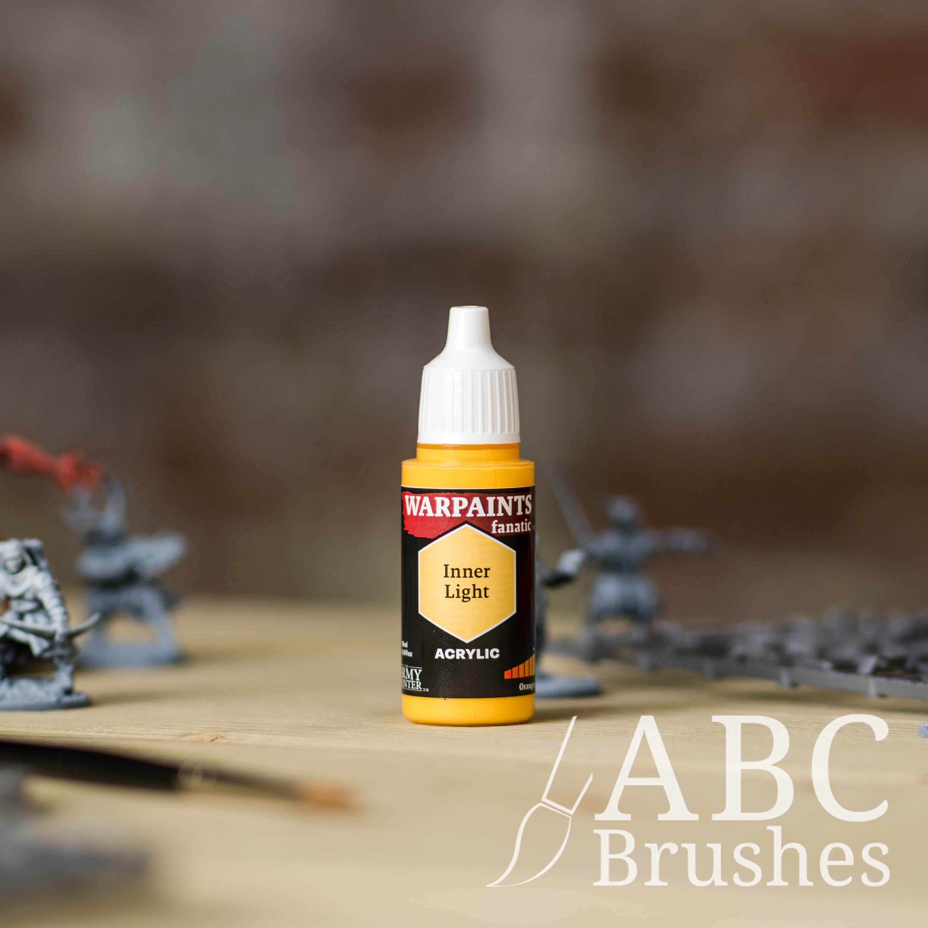 Bottle of the Army Painter Fanatics in the colour Inner Light with various wargaming models and an ABC paintbrush in the background, on a wooden surface.