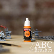 Bottle of the Army Painter Fanatics in the colour Lava Orange with various wargaming models and an ABC paintbrush in the background, on a wooden surface.
