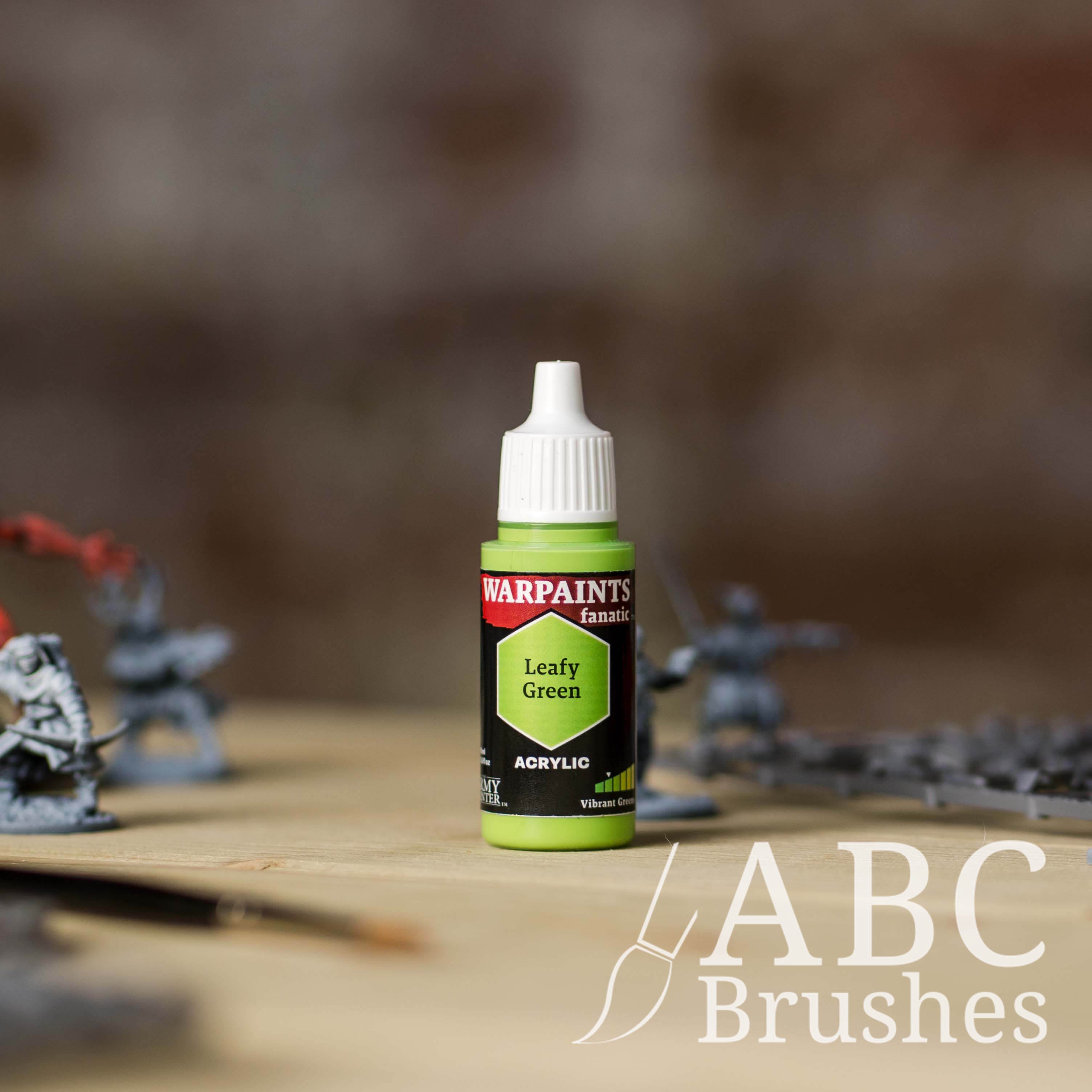 Bottle of the Army Painter Fanatics in the colour Leafy Green with various wargaming models and an ABC paintbrush in the background, on a wooden surface.