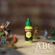 Bottle of the Army Painter Fanatics Effects in the colour Lens Flare Glow with various wargaming models and an ABC paintbrush in the background, on a wooden surface.