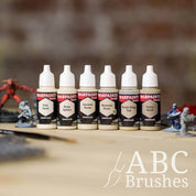 Army Painter Fanatics Light Neutrals Triad on a wooden surface. There are wargaming models and an ABC paint brush next to them.