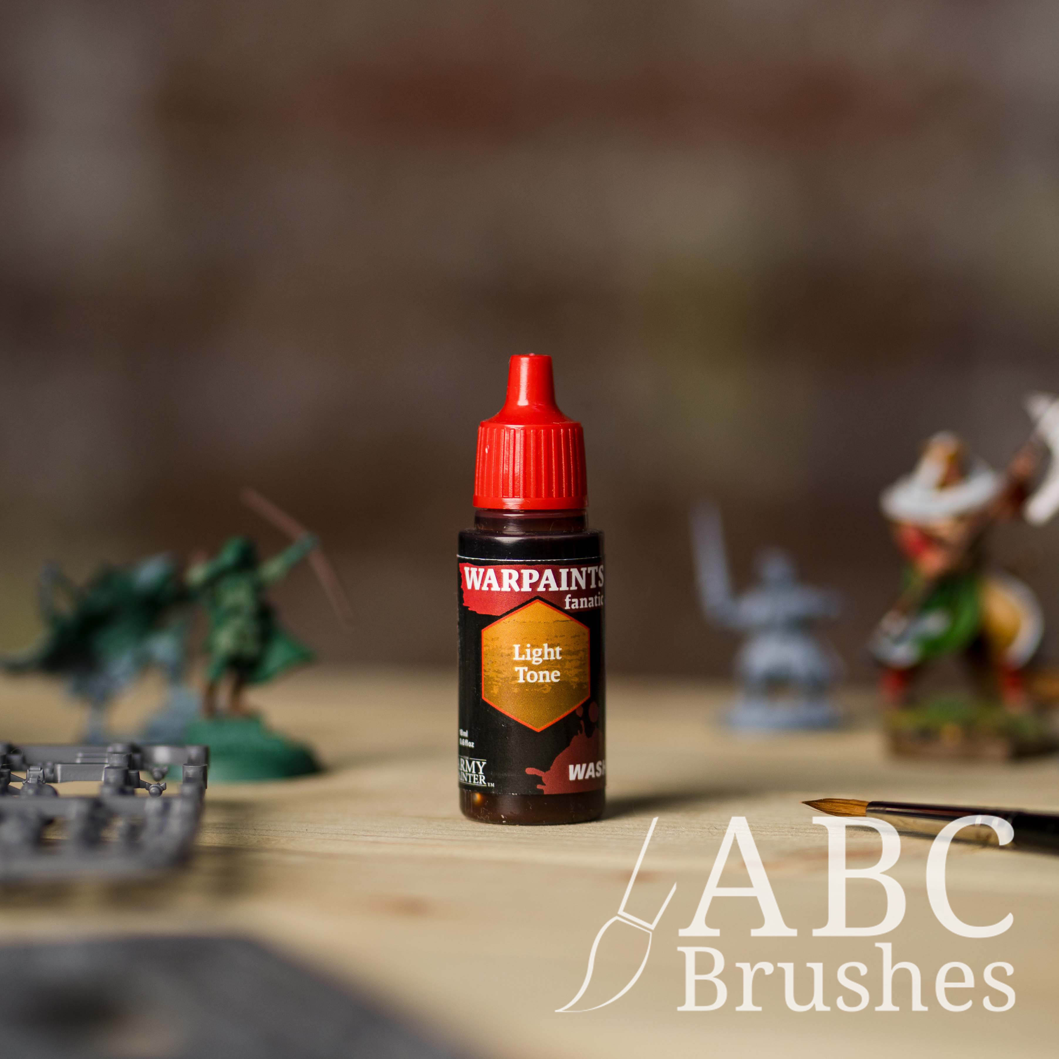 Bottle of the Army Painter Fanatics Wash in the colour Light Tone with various wargaming models and an ABC paintbrush in the background, on a wooden surface.