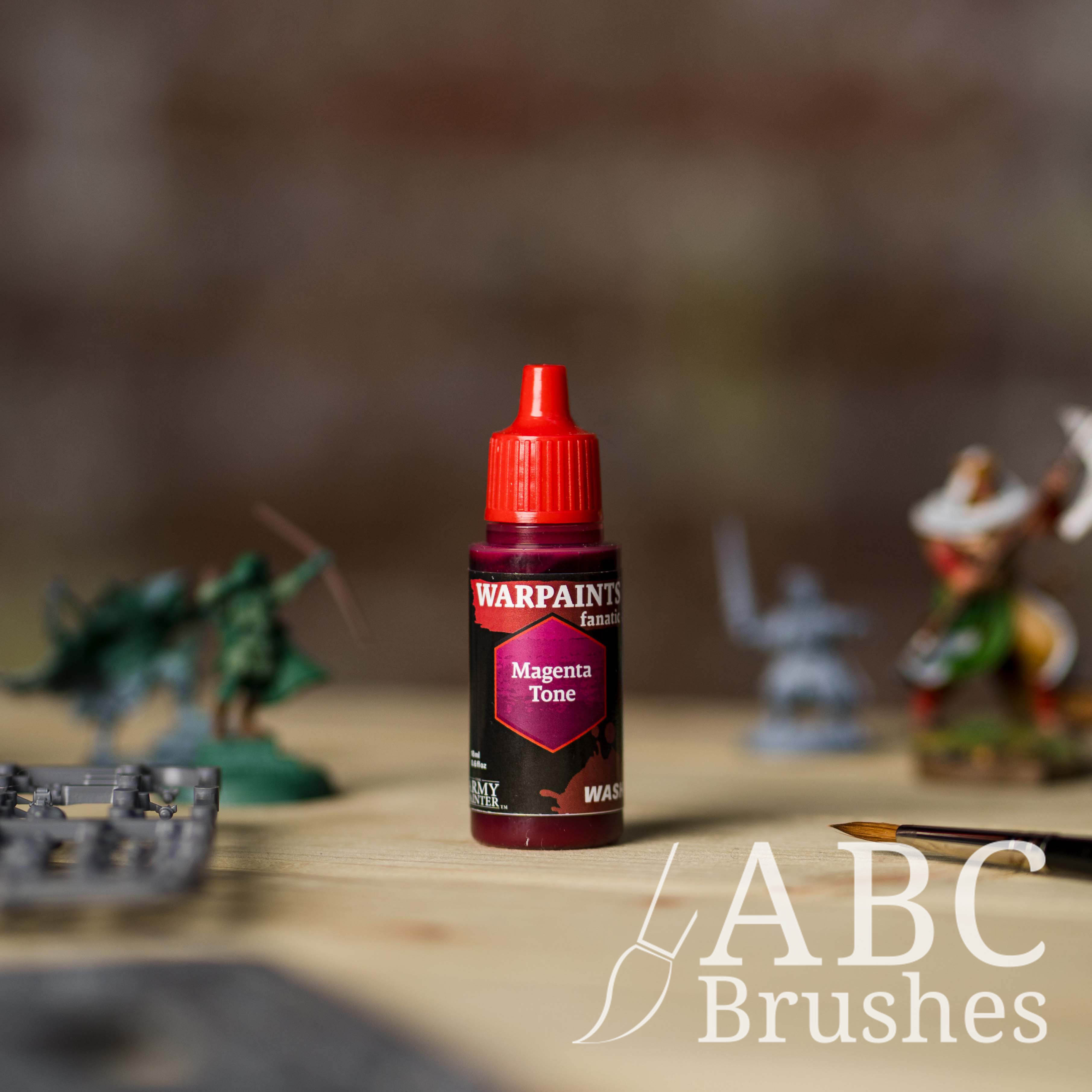 Bottle of the Army Painter Fanatics Wash in the colour Magenta Tone with various wargaming models and an ABC paintbrush in the background, on a wooden surface.