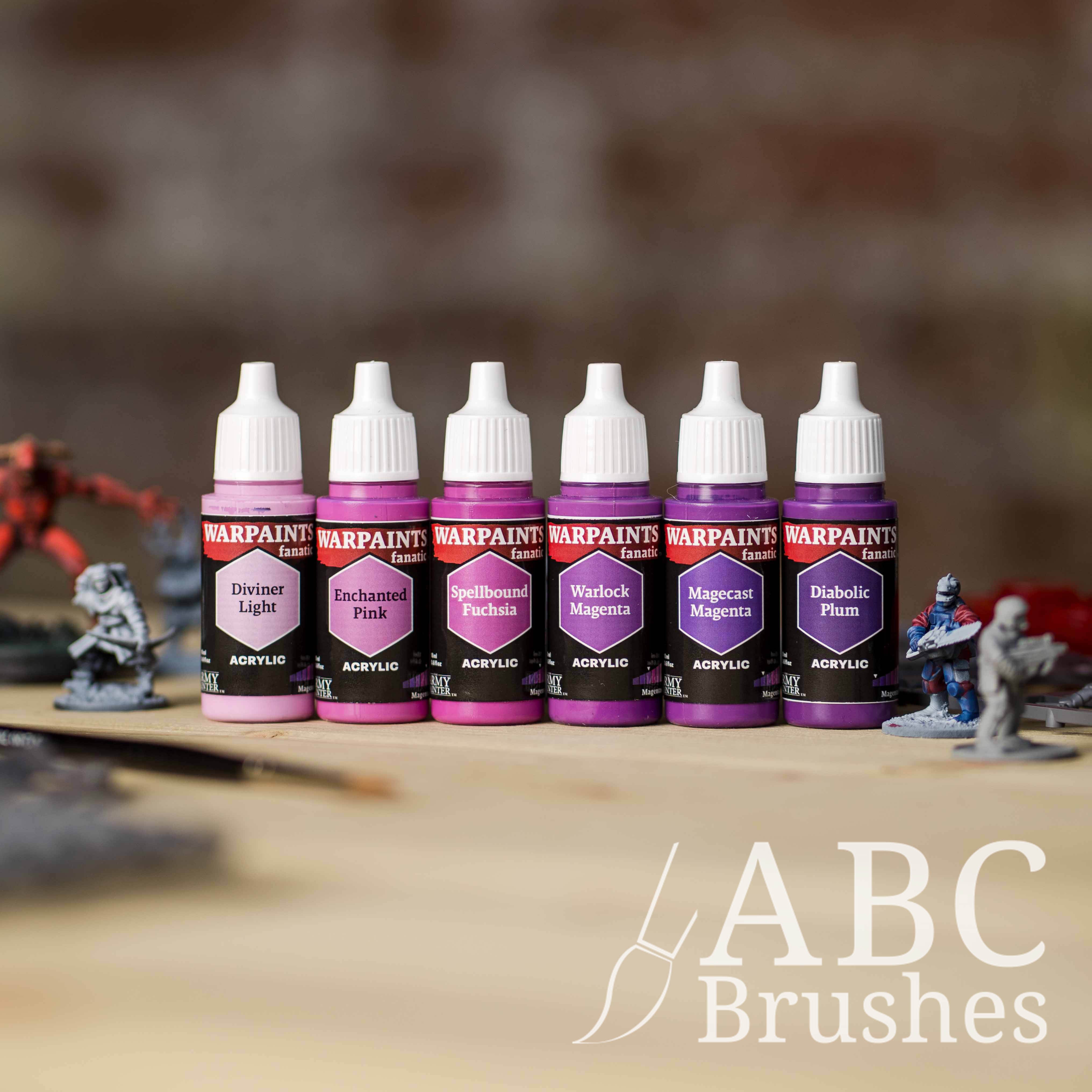 Army Painter Fanatics Magentas Triad on a wooden surface. There are wargaming models and an ABC paint brush next to them.