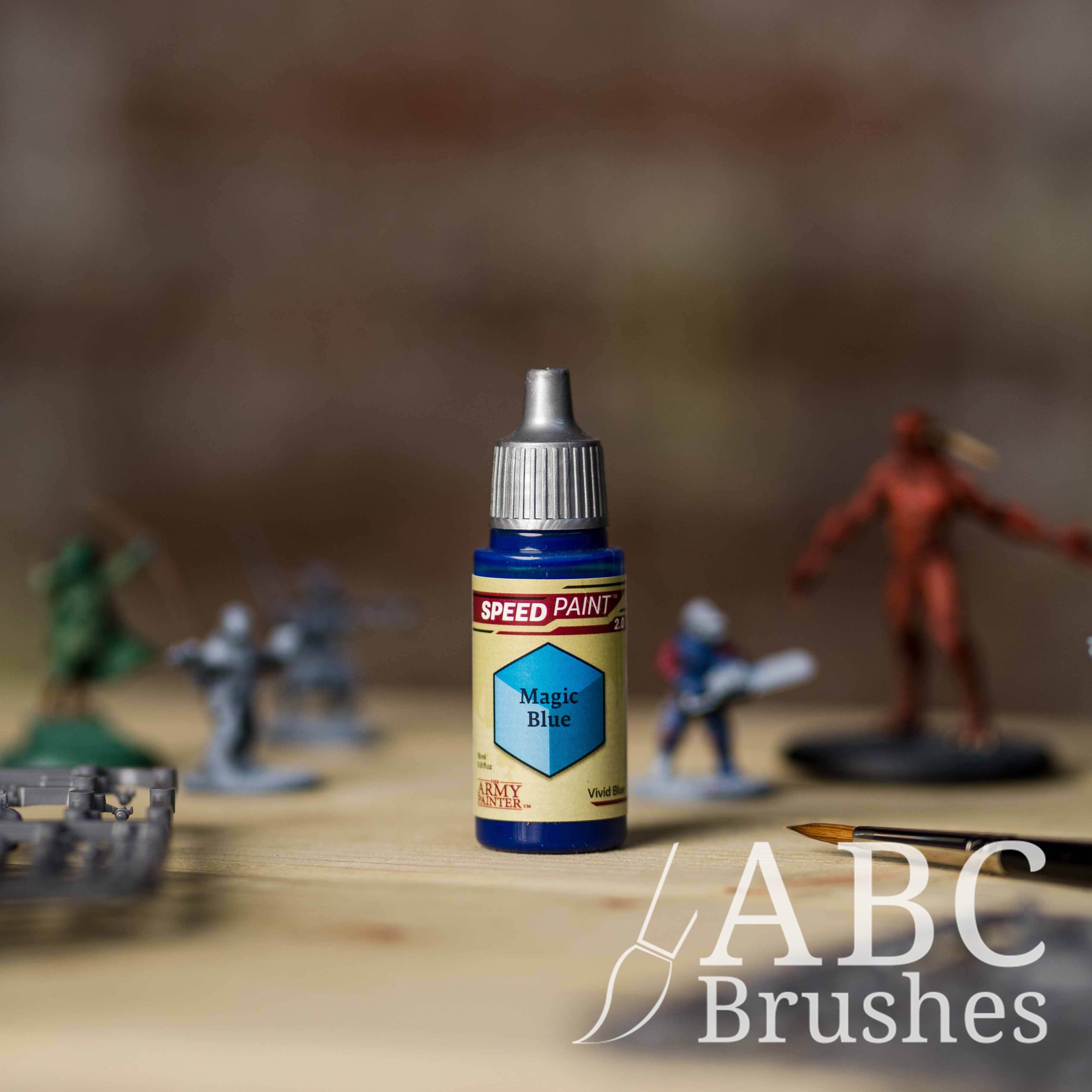 Bottle of the Army Painters Speedpaint in the colour Magic Blue, with wargaming models and an ABC paintbrush in the background, on a wooden surface.