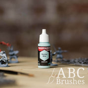 Bottle of the Army Painter Fanatics in the colour Marine Mist with various wargaming models and an ABC paintbrush in the background, on a wooden surface.