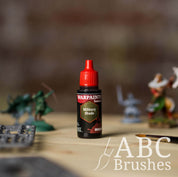 Bottle of the Army Painter Fanatics Wash in the colour Military Tone with various wargaming models and an ABC paintbrush in the background, on a wooden surface.