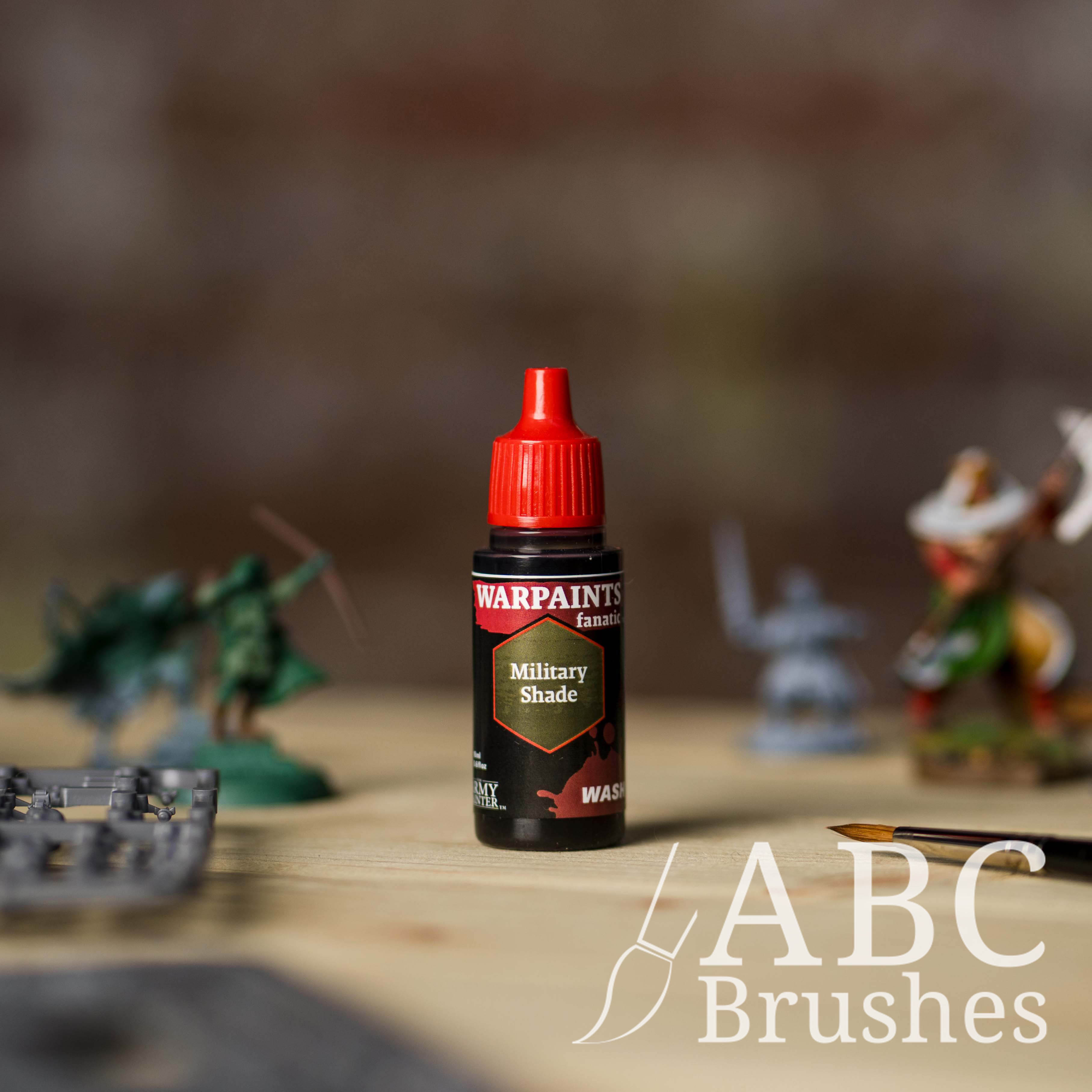 Bottle of the Army Painter Fanatics Wash in the colour Military Tone with various wargaming models and an ABC paintbrush in the background, on a wooden surface.