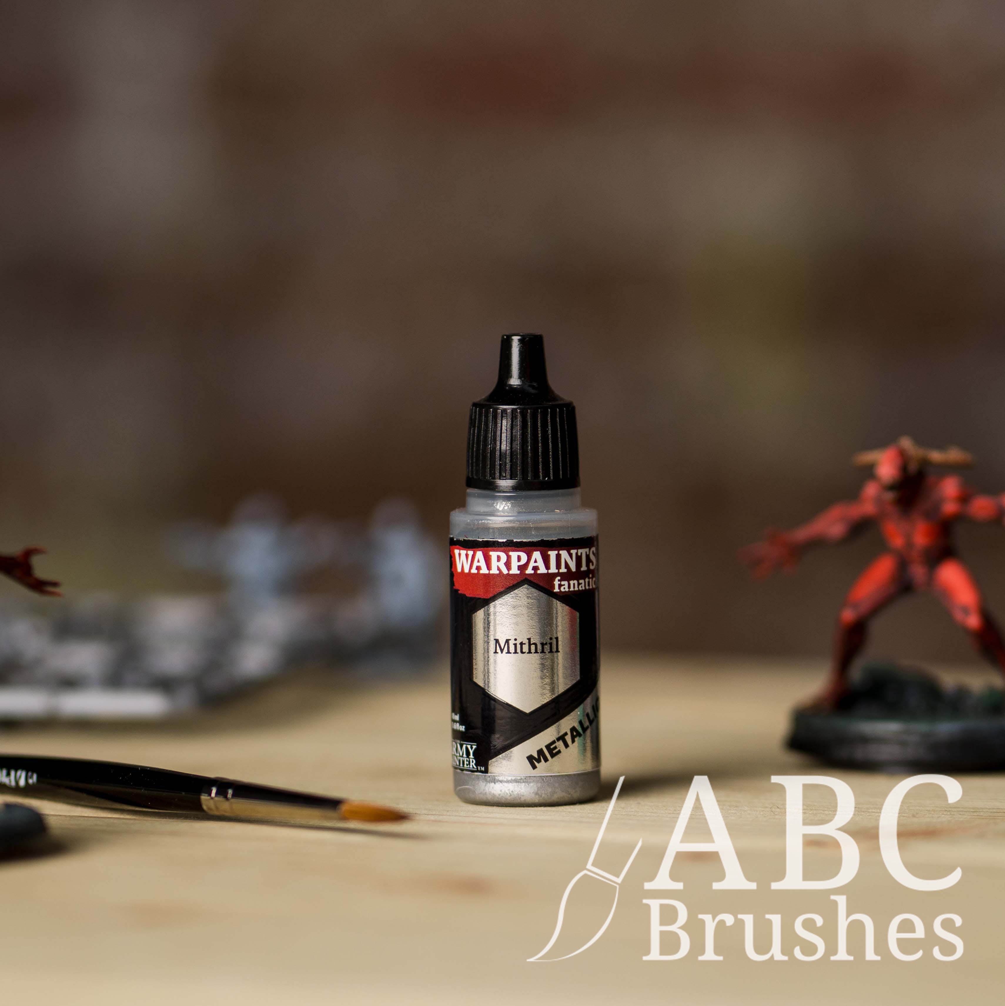 Bottle of the Army Painter Fanatics Metallic paint in the colour Mithril with various wargaming models and an ABC paintbrush in the background, on a wooden surface.