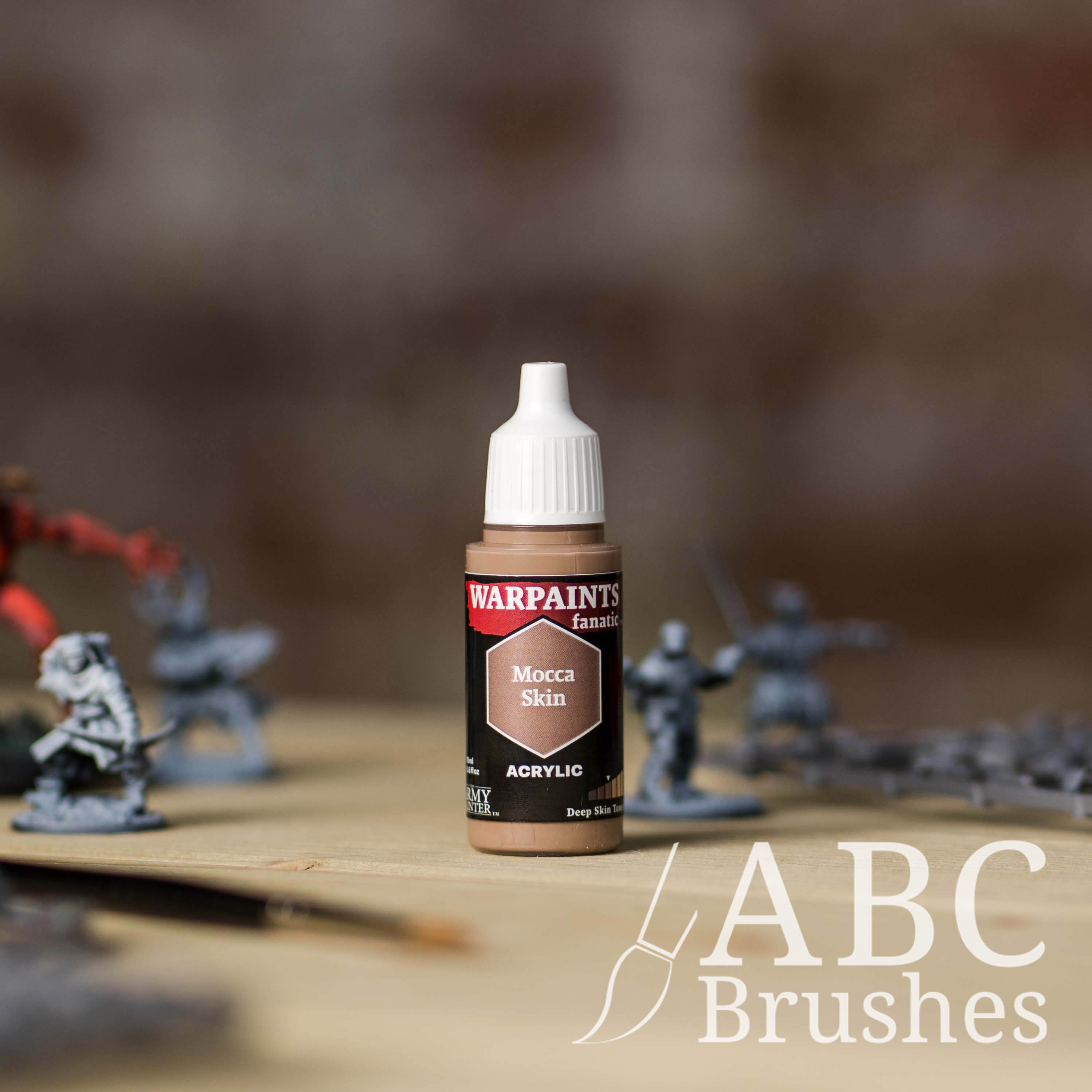 Bottle of the Army Painter Fanatics in the colour Mocca Skin with various wargaming models and an ABC paintbrush in the background, on a wooden surface.