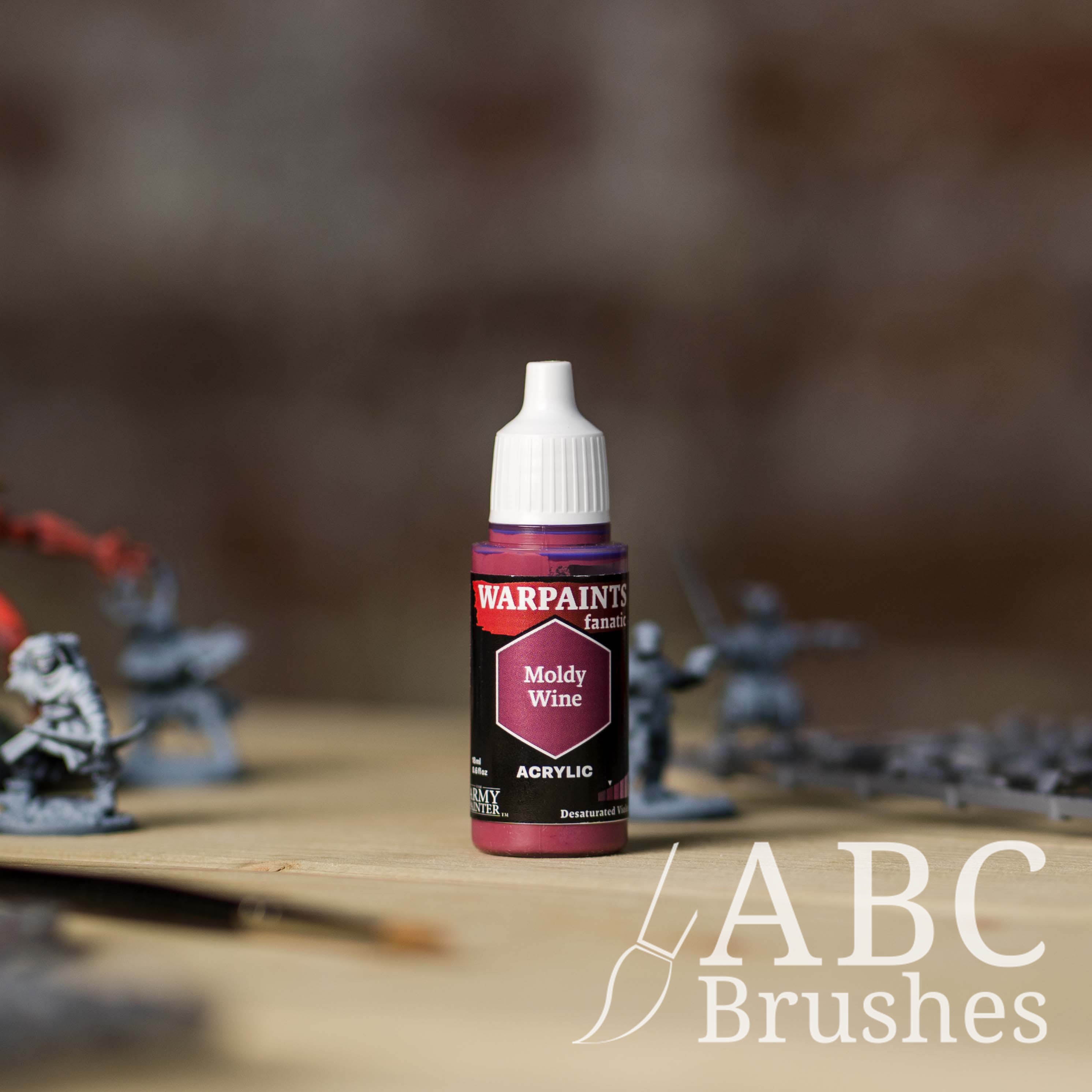 Bottle of the Army Painter Fanatics in the colour Moldy Wine with various wargaming models and an ABC paintbrush in the background, on a wooden surface.