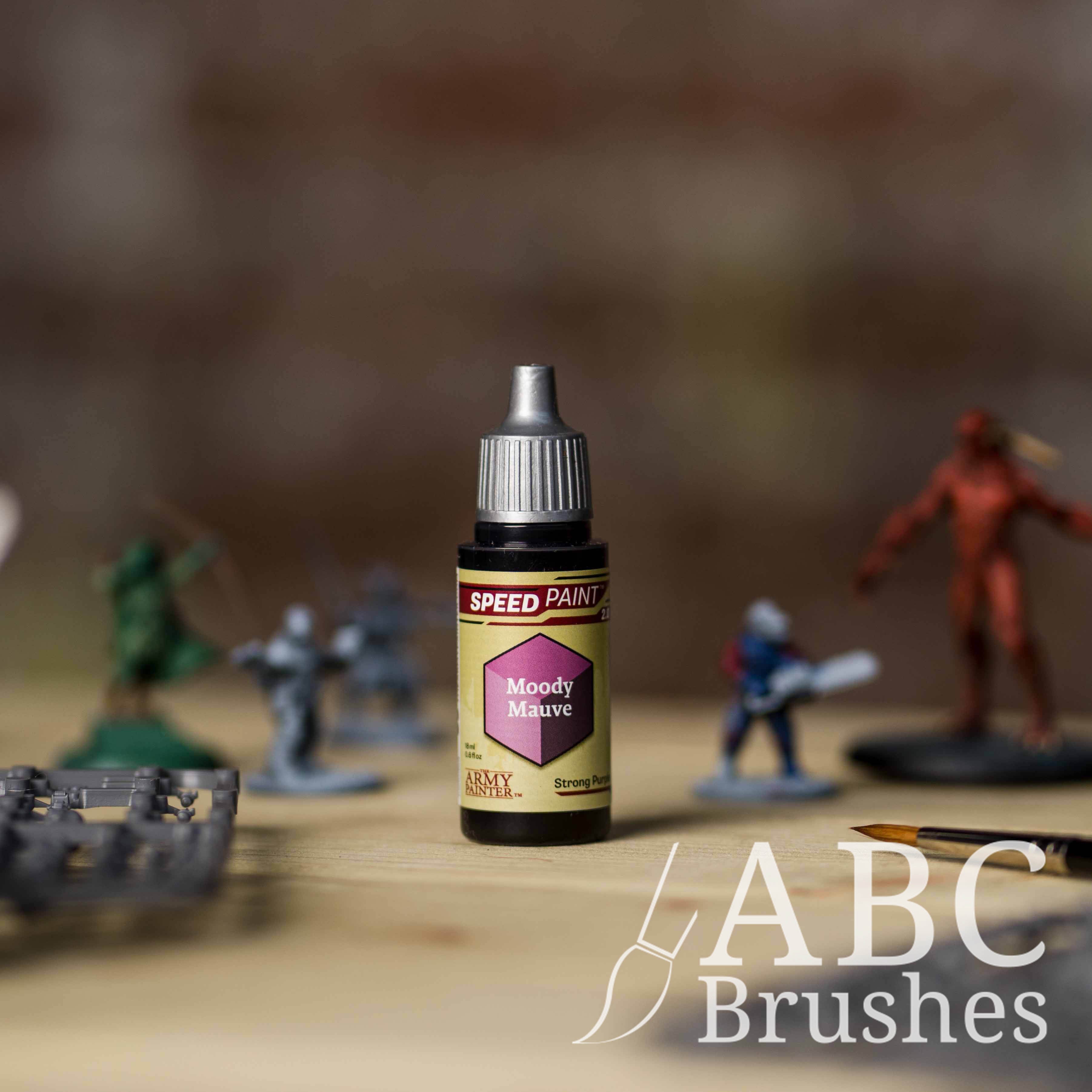 Bottle of the Army Painters Speedpaint in the colour Moody Mauve, with wargaming models and an ABC paintbrush in the background, on a wooden surface.