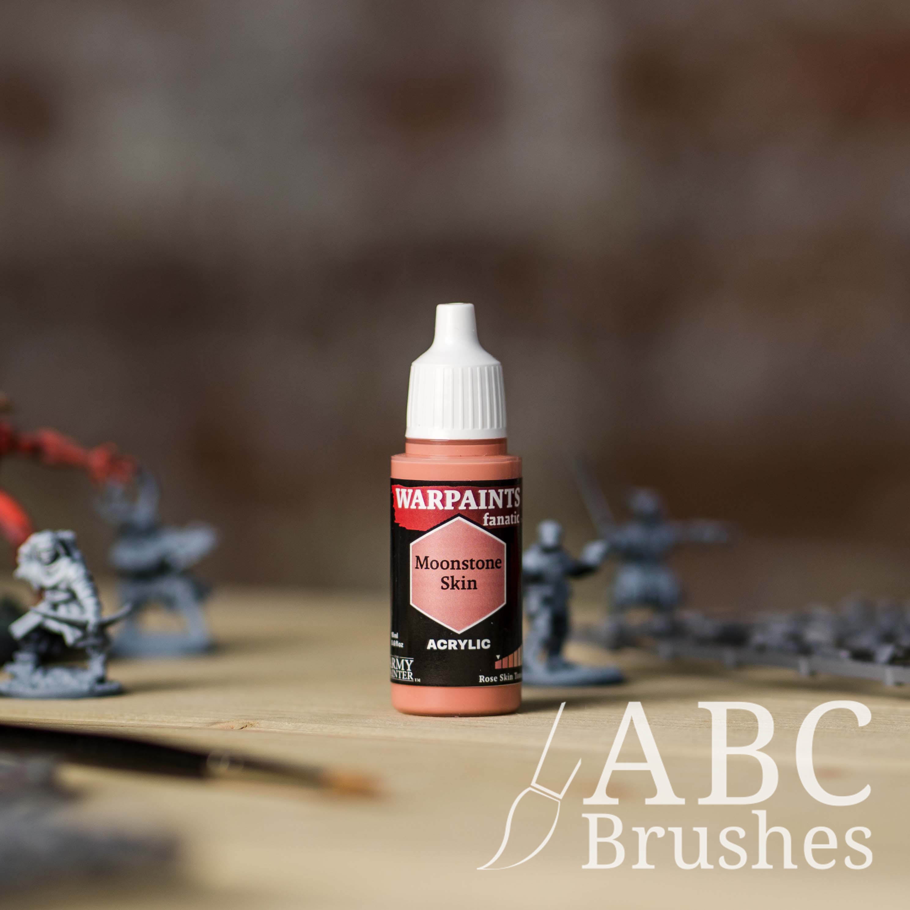 Bottle of the Army Painter Fanatics in the colour Moonstone Skin with various wargaming models and an ABC paintbrush in the background, on a wooden surface.