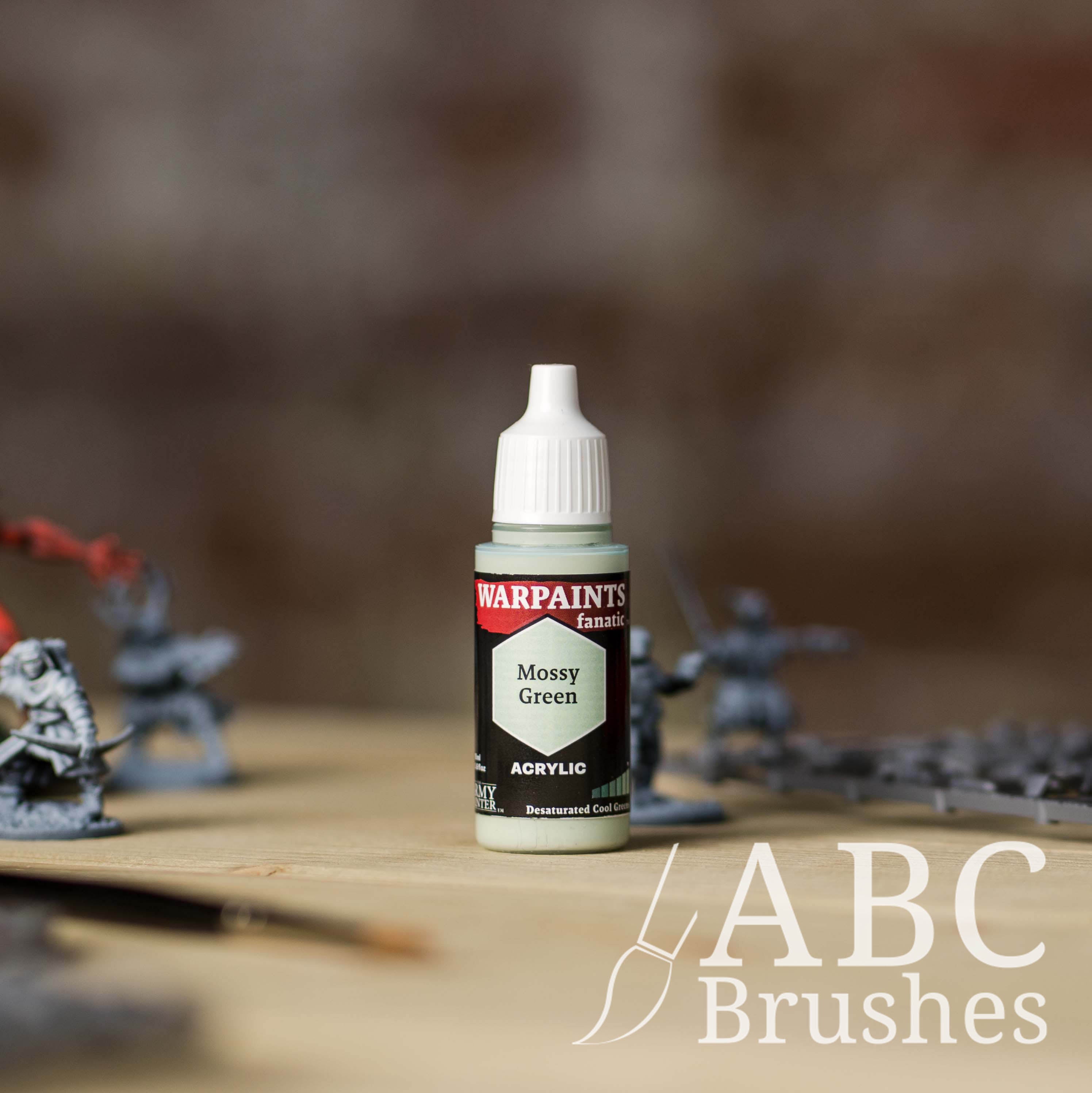 Bottle of the Army Painter Fanatics in the colour Mossy Green with various wargaming models and an ABC paintbrush in the background, on a wooden surface.