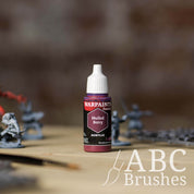 Bottle of the Army Painter Fanatics in the colour Mulled Berry with various wargaming models and an ABC paintbrush in the background, on a wooden surface.