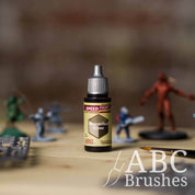 Bottle of the Army Painters Speedpaint in the colour Mummified Grime, with wargaming models and an ABC paintbrush in the background, on a wooden surface.