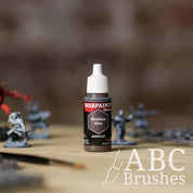 Bottle of the Army Painter Fanatics in the colour Obsidian Skin with various wargaming models and an ABC paintbrush in the background, on a wooden surface.