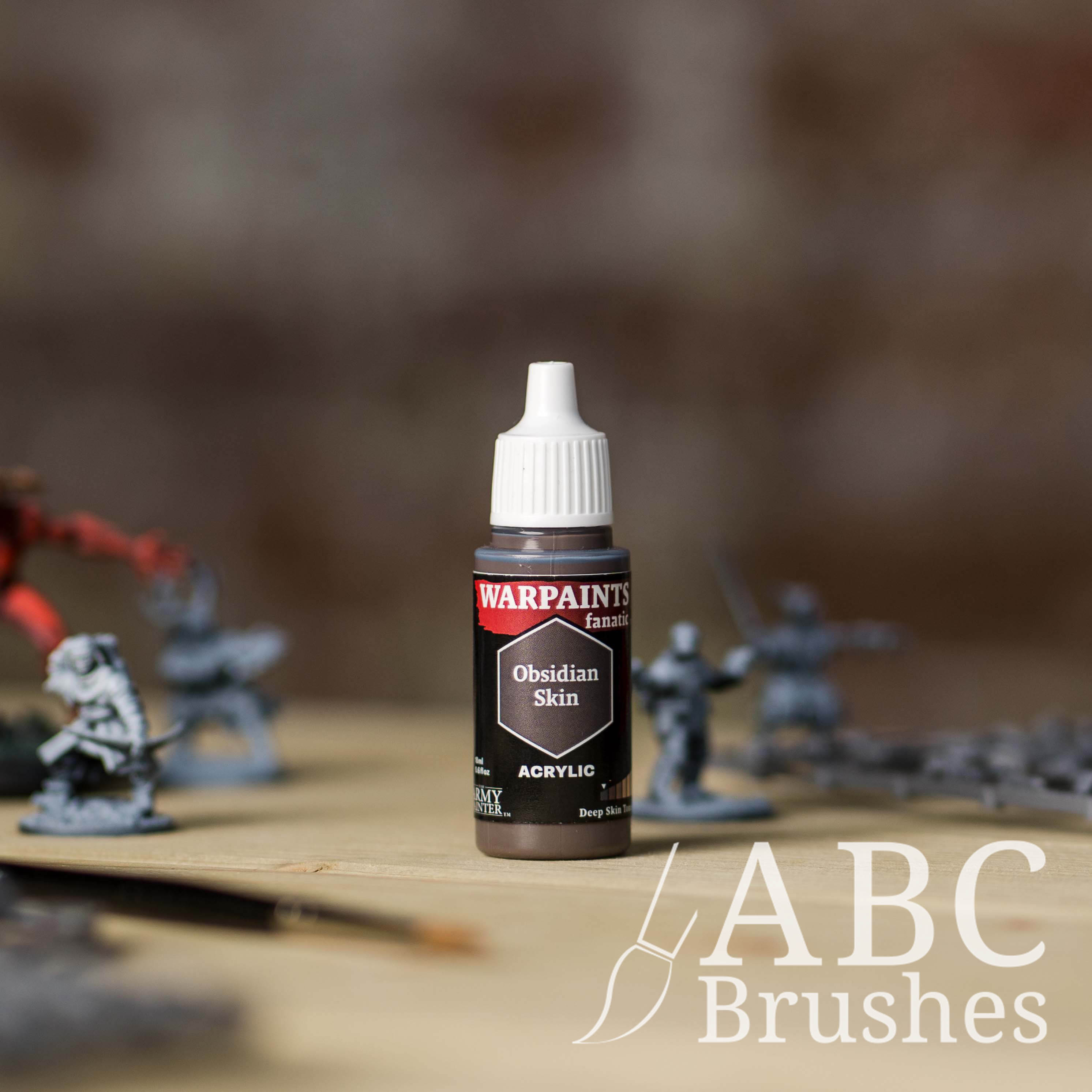 Bottle of the Army Painter Fanatics in the colour Obsidian Skin with various wargaming models and an ABC paintbrush in the background, on a wooden surface.