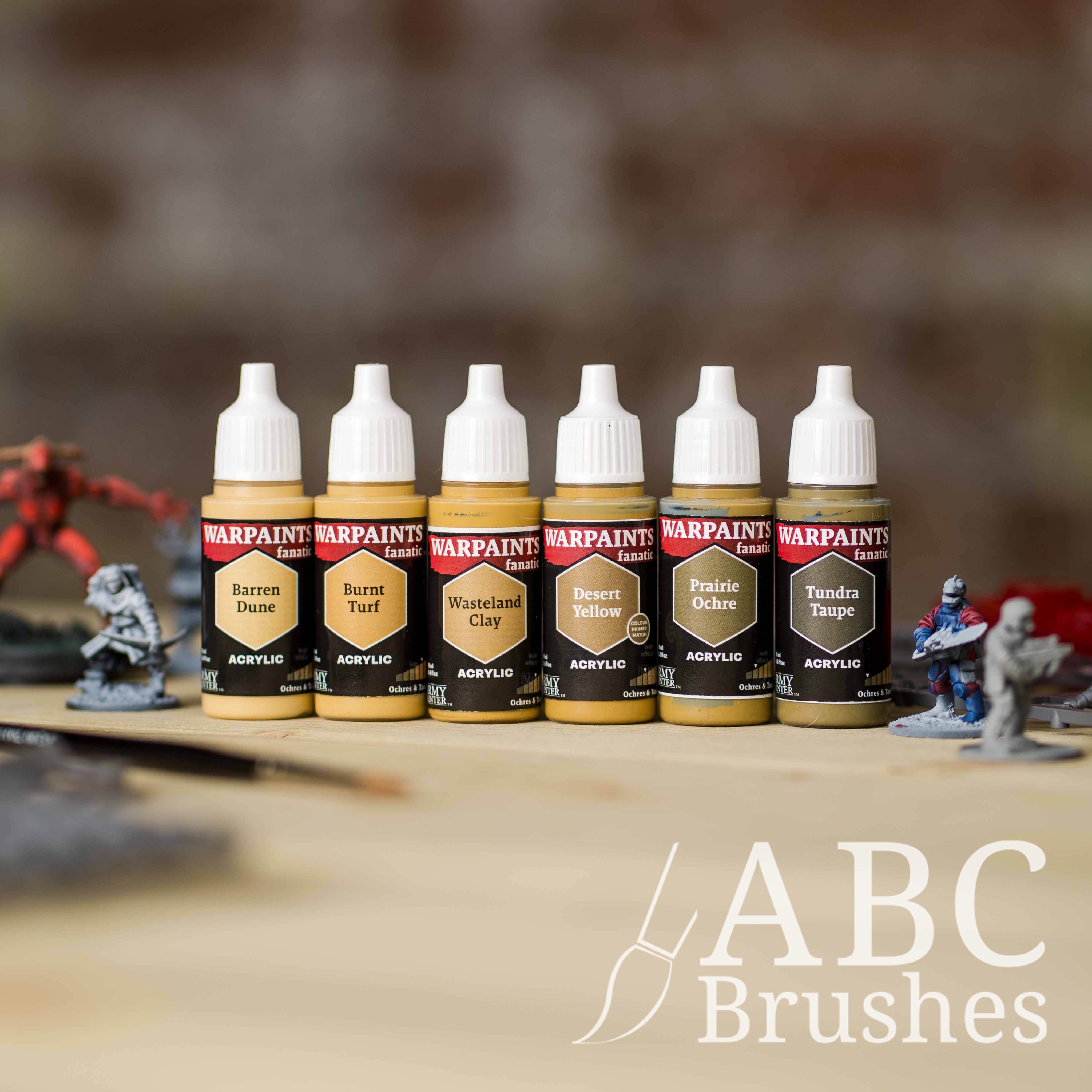Army Painter Fanatics Ochres and Tans Triad on a wooden surface. There are wargaming models and an ABC paint brush next to them.