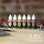 Army Painter Fanatics Olive Green Triad on a wooden surface. There are wargaming models and an ABC paint brush next to them.