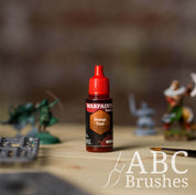 Bottle of the Army Painter Fanatics Wash in the colour Orange Tone with various wargaming models and an ABC paintbrush in the background, on a wooden surface.