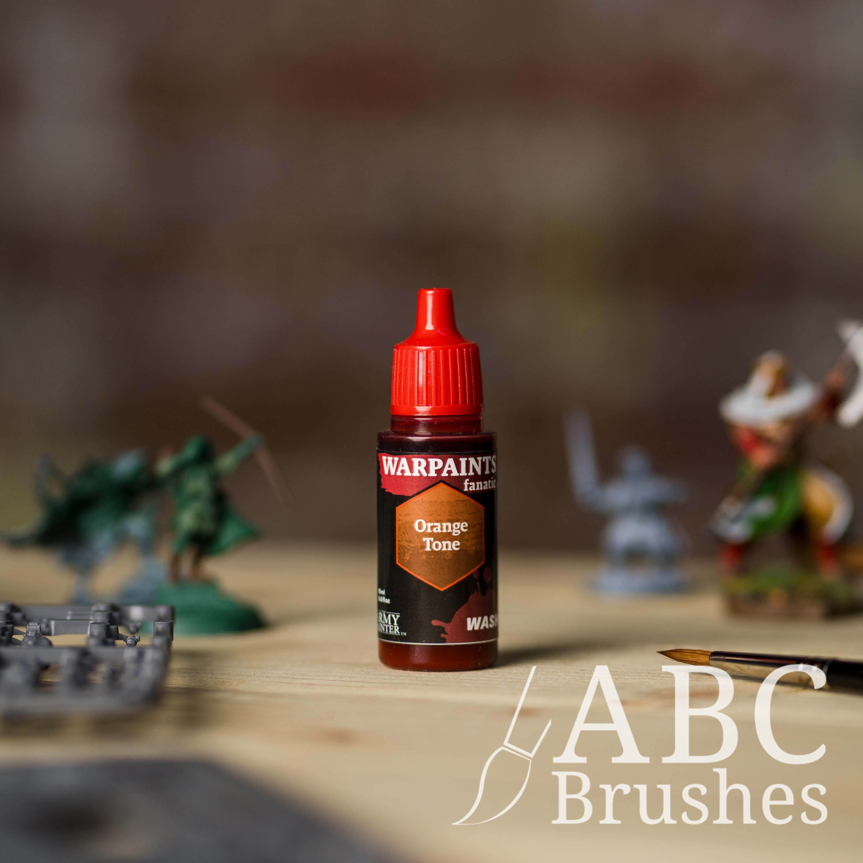 Bottle of the Army Painter Fanatics Wash in the colour Orange Tone with various wargaming models and an ABC paintbrush in the background, on a wooden surface.