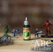 Bottle of the Army Painters Speedpaint in the colour Orc Skin, with wargaming models and an ABC paintbrush in the background, on a wooden surface.