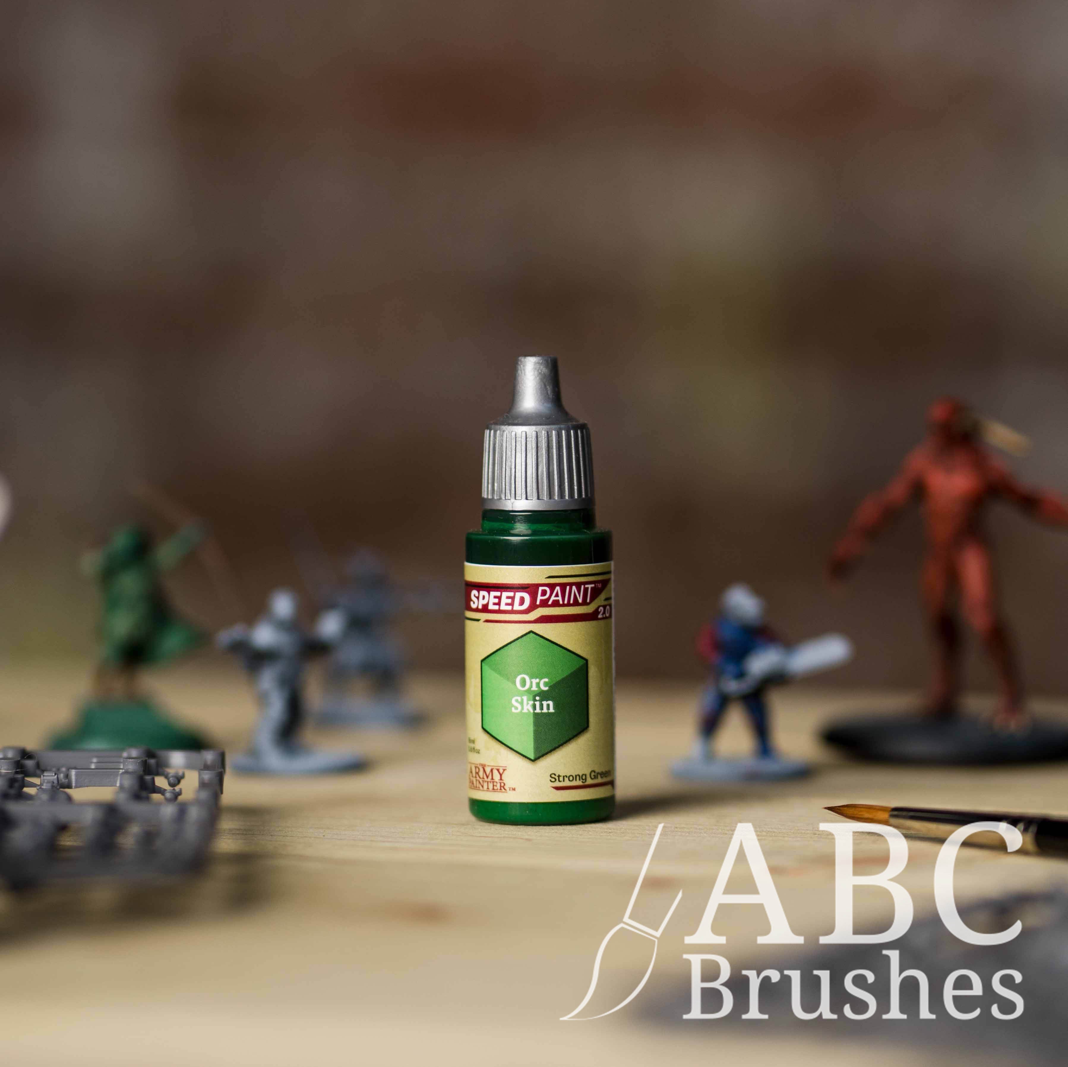 Bottle of the Army Painters Speedpaint in the colour Orc Skin, with wargaming models and an ABC paintbrush in the background, on a wooden surface.