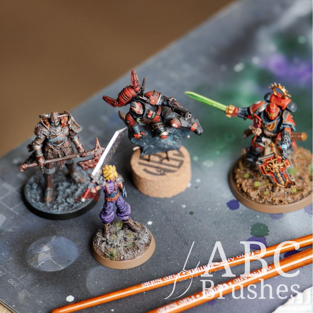 Four wargaming miniatures on a paint covered cutting mat, next to two ABC paintbrushes. 