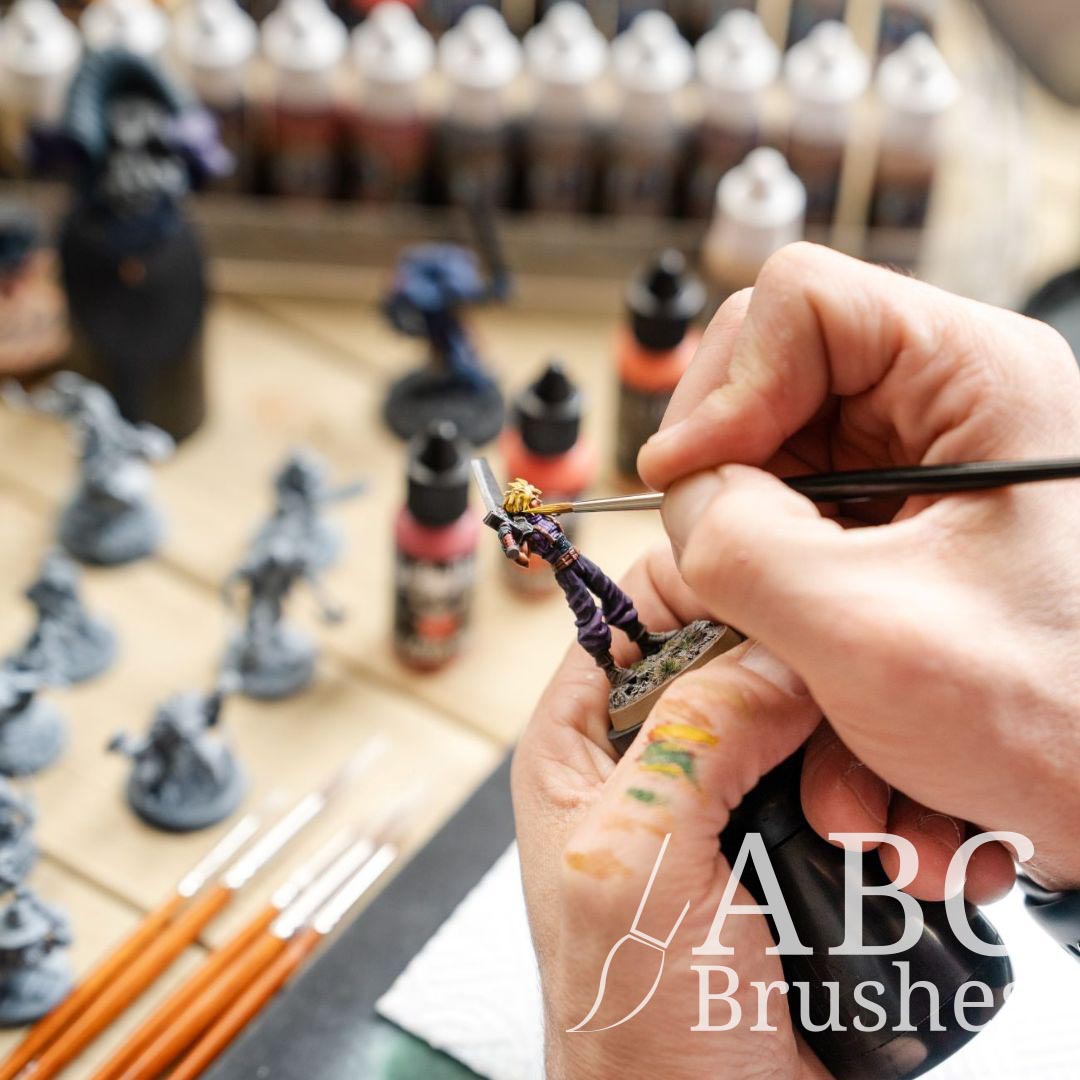 Painting a mini figure with an ABC paint brush. There is an array of acrylic paint bottles and other mini figures in the background. 