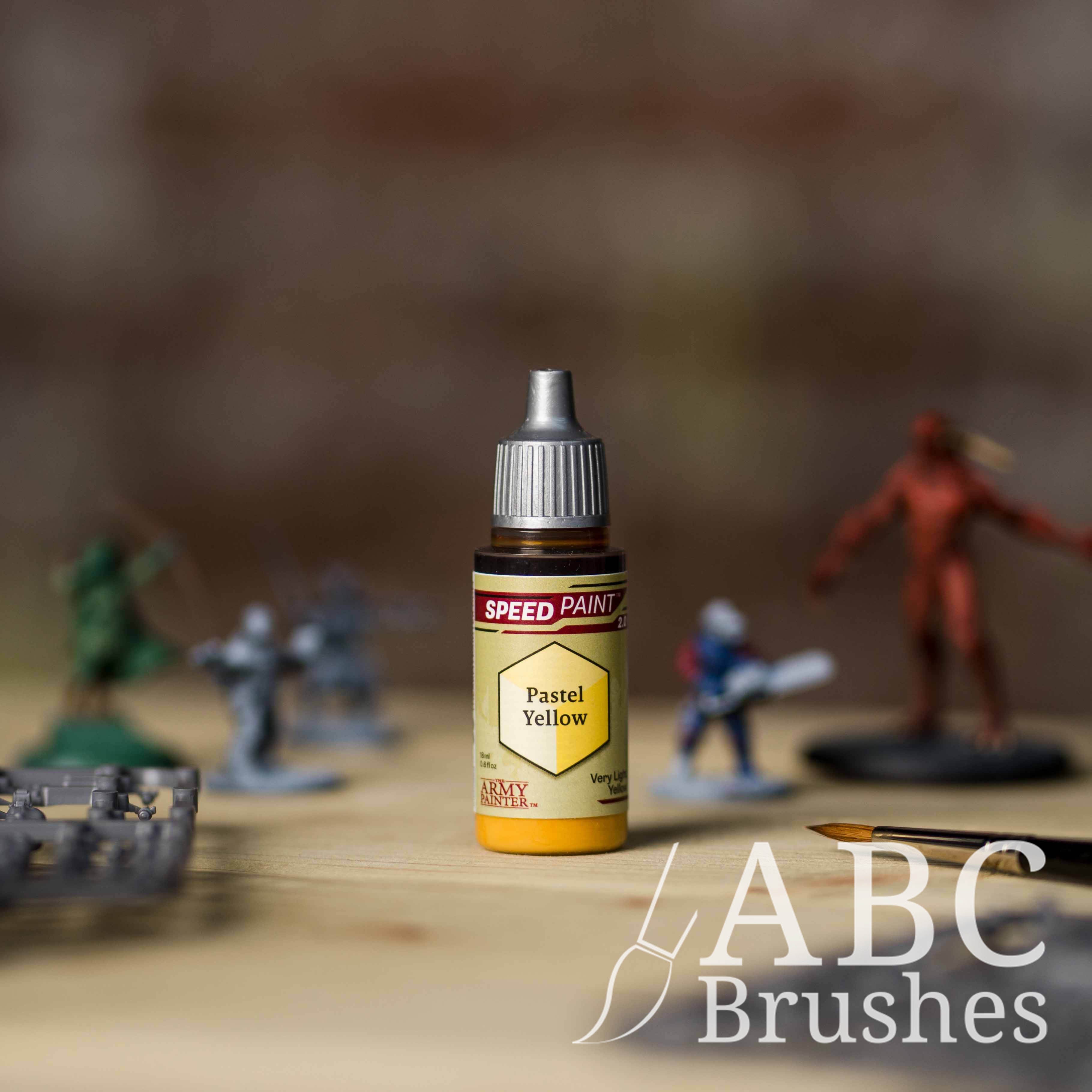 Bottle of the Army Painters Speedpaint in the colour Pastel Yellow, with wargaming models and an ABC paintbrush in the background, on a wooden surface.