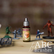 Bottle of the Army Painters Speedpaint in the colour Peachy Flesh, with wargaming models and an ABC paintbrush in the background, on a wooden surface.
