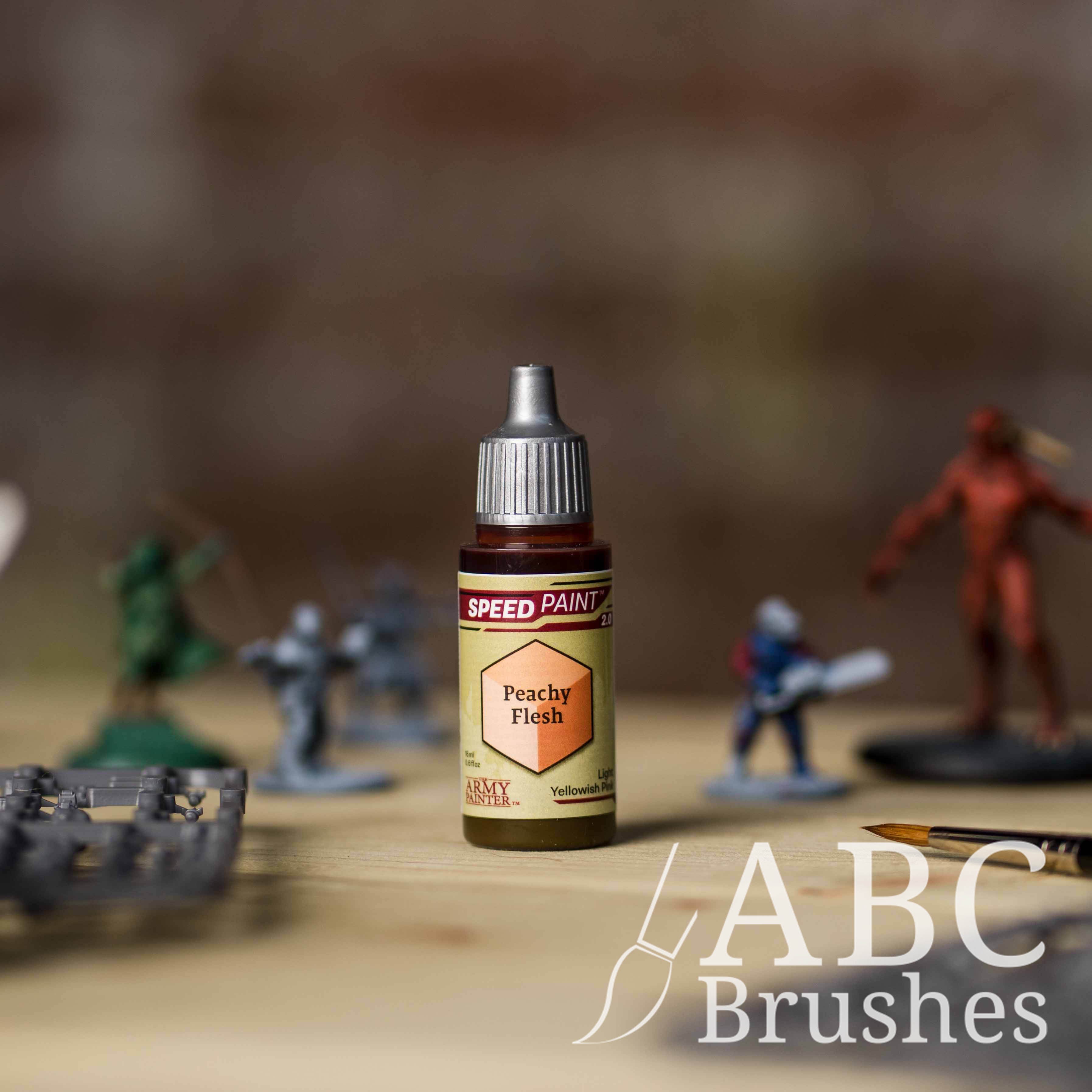 Bottle of the Army Painters Speedpaint in the colour Peachy Flesh, with wargaming models and an ABC paintbrush in the background, on a wooden surface.